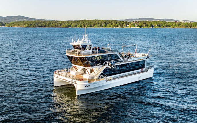 Dinner cruise boat on Oslofjord, Norway, with scenic forested shoreline in the background.