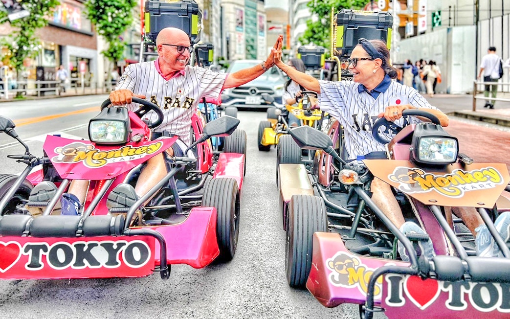 Two men in Tokyo jerseys high-five while driving go-karts on a city street.
