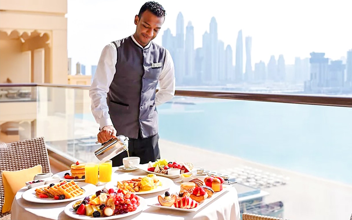 Server pouring coffee on a balcony with breakfast spread at Fairmont Hotel, Dubai skyline in background.