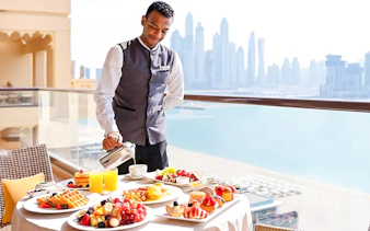 Server pouring coffee on a balcony with breakfast spread at Fairmont Hotel, Dubai skyline in background.