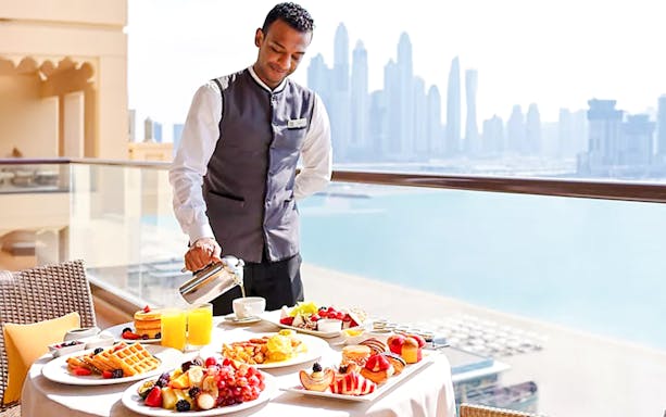Server pouring coffee on a balcony with breakfast spread at Fairmont Hotel, Dubai skyline in background.
