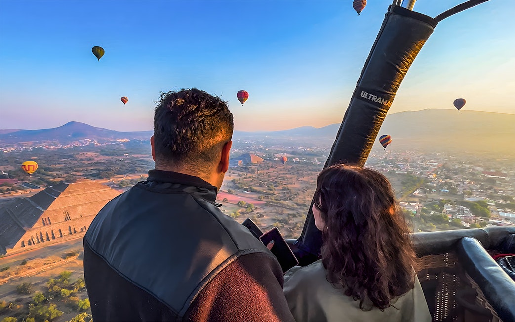 Passengers in hot air balloon over Teotihuacan pyramid, Mexico, with other balloons in the sky.