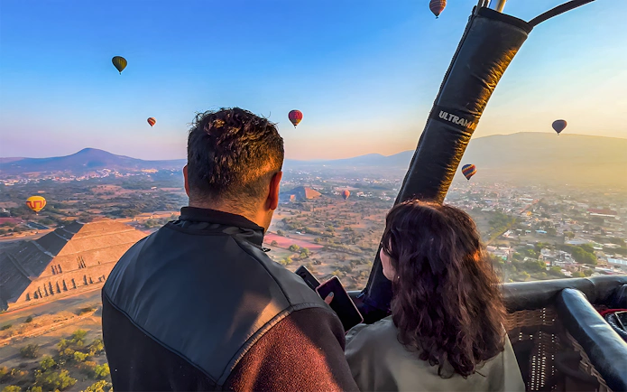 Passengers in hot air balloon over Teotihuacan pyramid, Mexico, with other balloons in the sky.