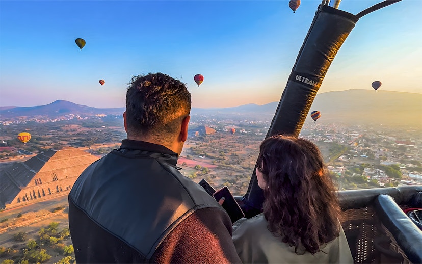 Passengers in hot air balloon over Teotihuacan pyramid, Mexico, with other balloons in the sky.