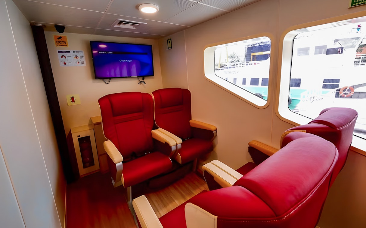 Red seating area inside Sindo Ferry at Tanah Merah Terminal with window view of docked ship.