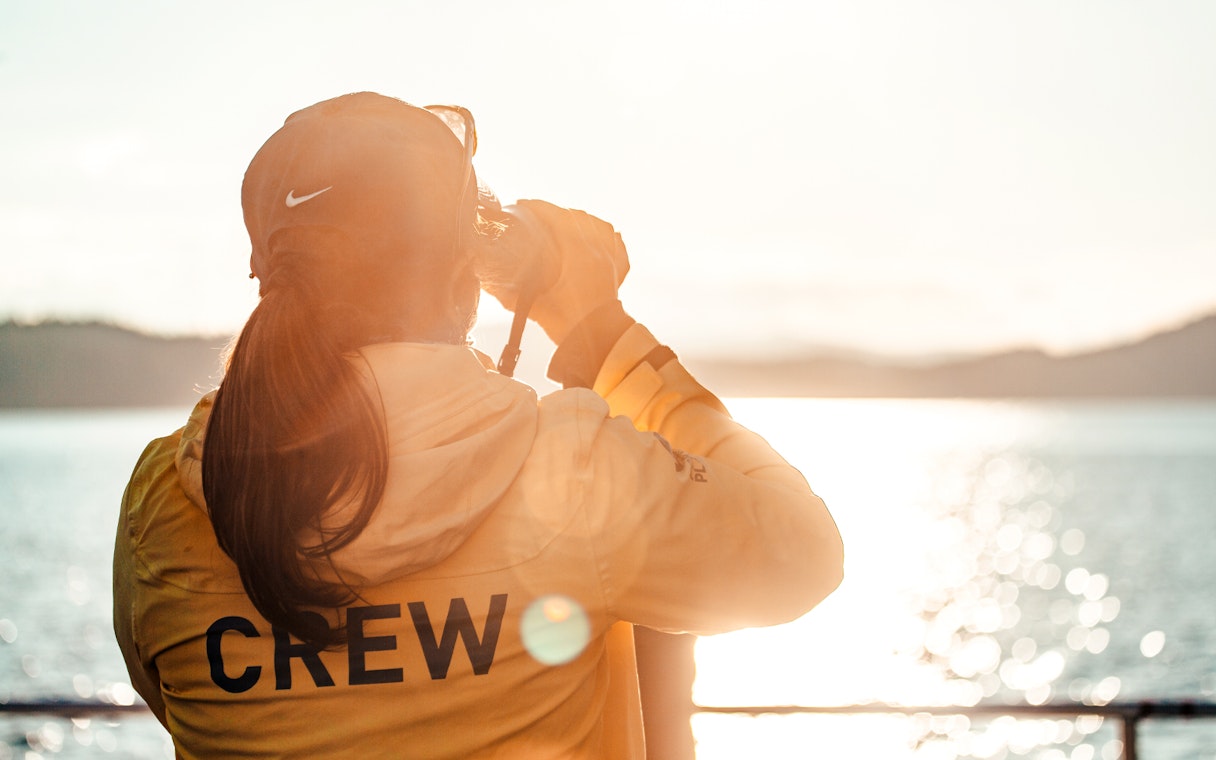 Person in yellow jacket with "CREW" on back, looking through binoculars over a sunlit lake.