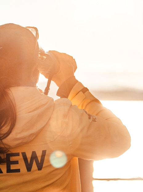 Person in yellow jacket with "CREW" on back, looking through binoculars over a sunlit lake.