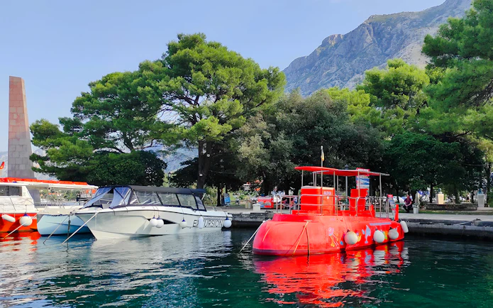Docked red semi-submarine in Boka Bay, Kotor, with mountains and trees in the background.