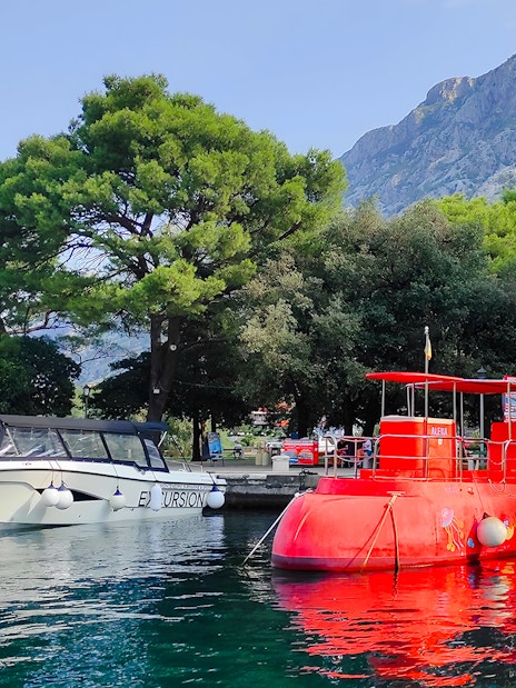 Docked red semi-submarine in Boka Bay, Kotor, with mountains and trees in the background.
