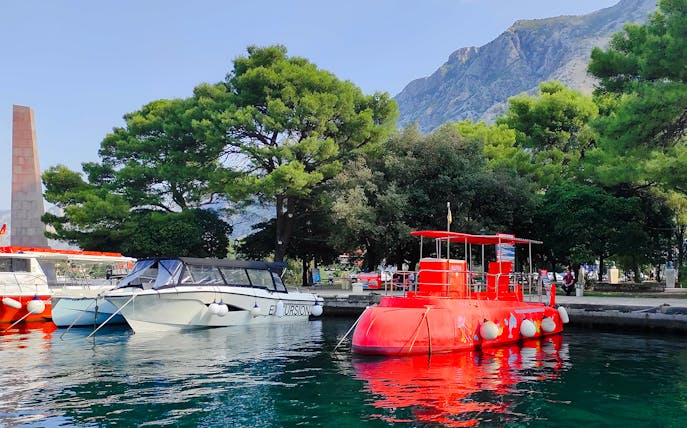 Docked red semi-submarine in Boka Bay, Kotor, with mountains and trees in the background.