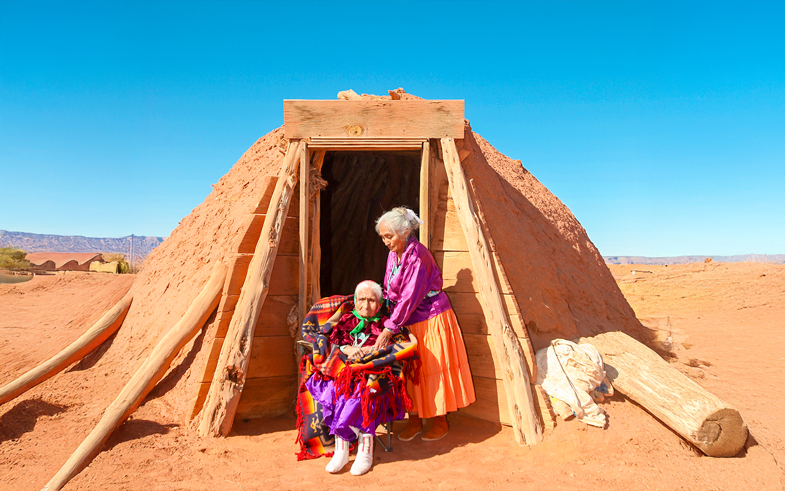 Navajo women in traditional attire outside a Hogan hut in a desert landscape.