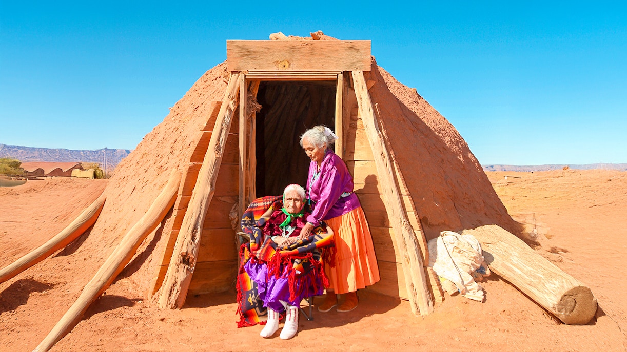 Navajo women in traditional attire outside a Hogan hut in a desert landscape.