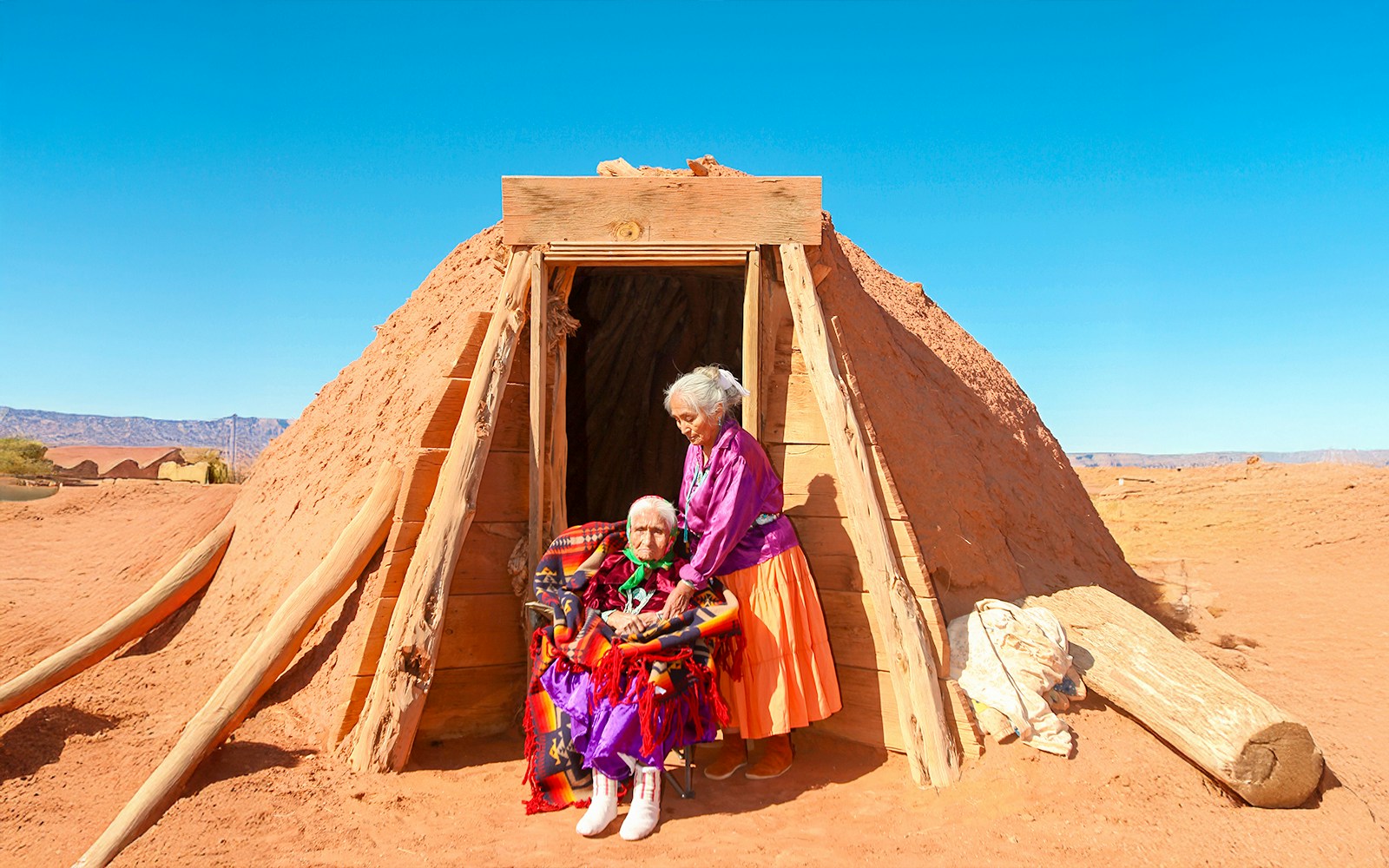Navajo women in traditional attire outside a Hogan hut in a desert landscape.