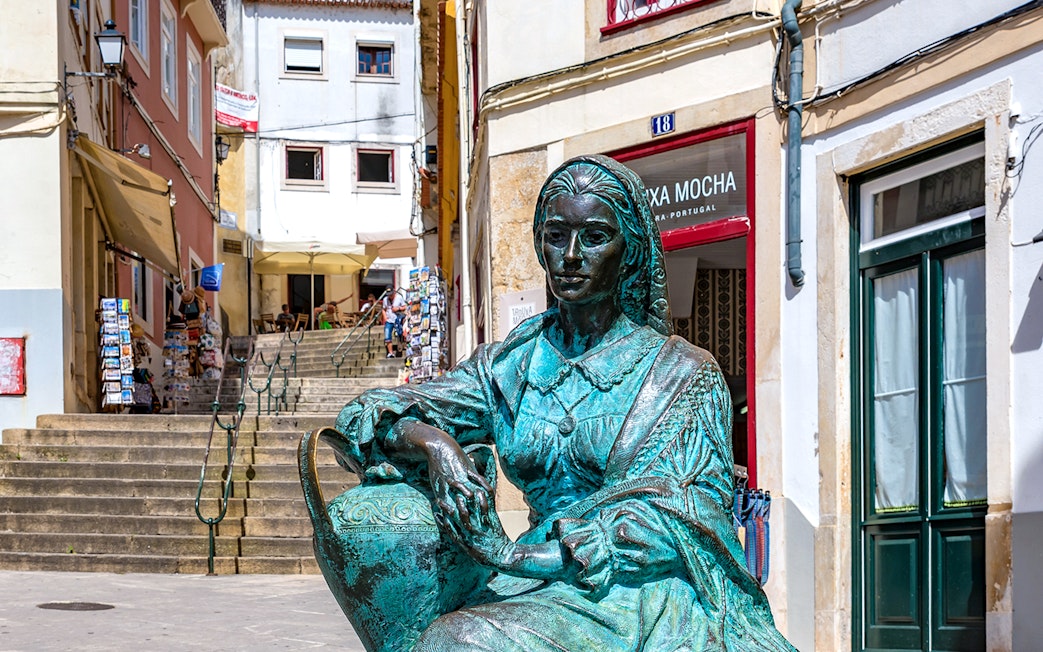 Bronze statue of a woman in Coimbra, Portugal, near a staircase with shops.