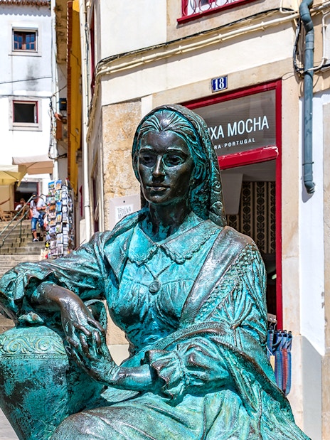 Bronze statue of a woman in Coimbra, Portugal, near a staircase with shops.