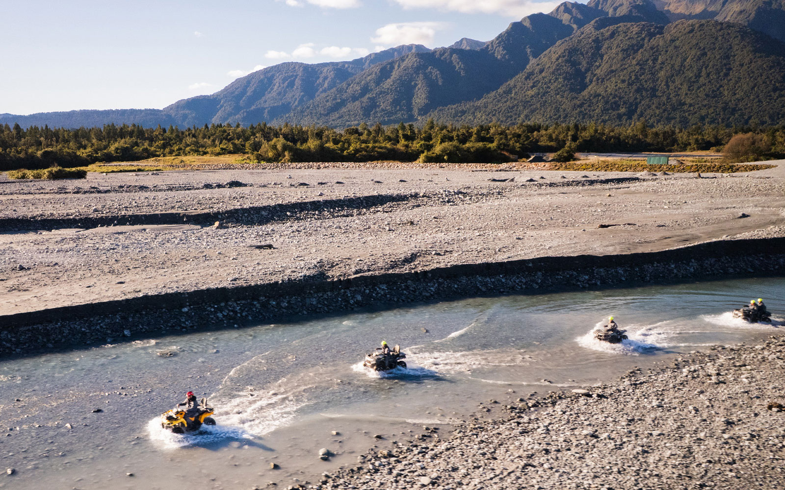 Quad biking through riverbed at Franz Josef, New Zealand with mountain backdrop.