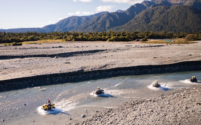Quad biking through riverbed at Franz Josef, New Zealand with mountain backdrop.