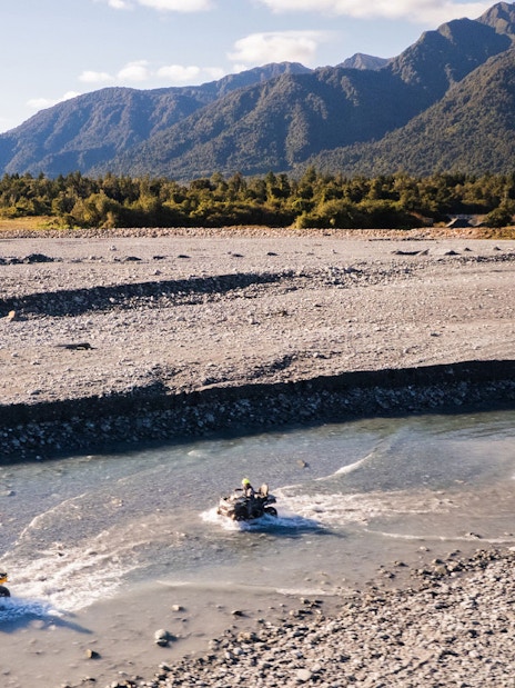 Quad biking through riverbed at Franz Josef, New Zealand with mountain backdrop.