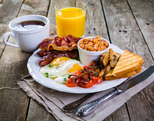English breakfast with eggs, bacon, sausages, beans, mushrooms, tomatoes, toast, tea, and orange juice.