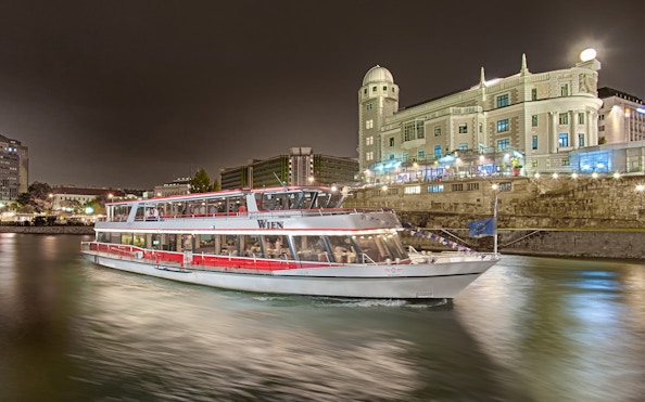 Cruise boat on the Danube River at night with illuminated historic building in Vienna.