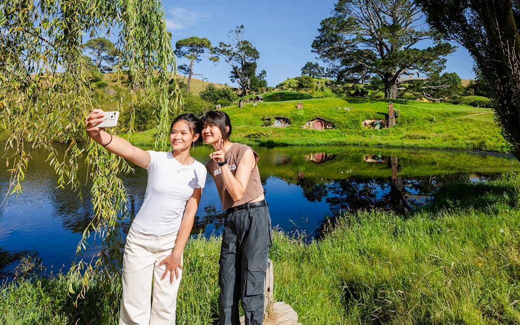 Girls taking a selfie with Hobbiton movie set hobbit houses in the background.