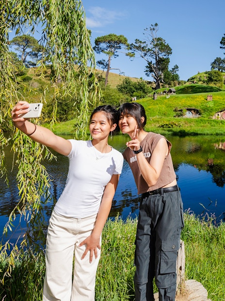 Girls taking a selfie with Hobbiton movie set hobbit houses in the background.