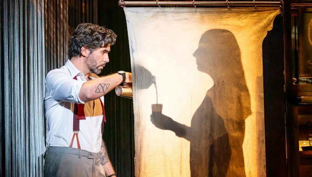 Man pouring drink with shadow silhouette on curtain during theatrical performance.