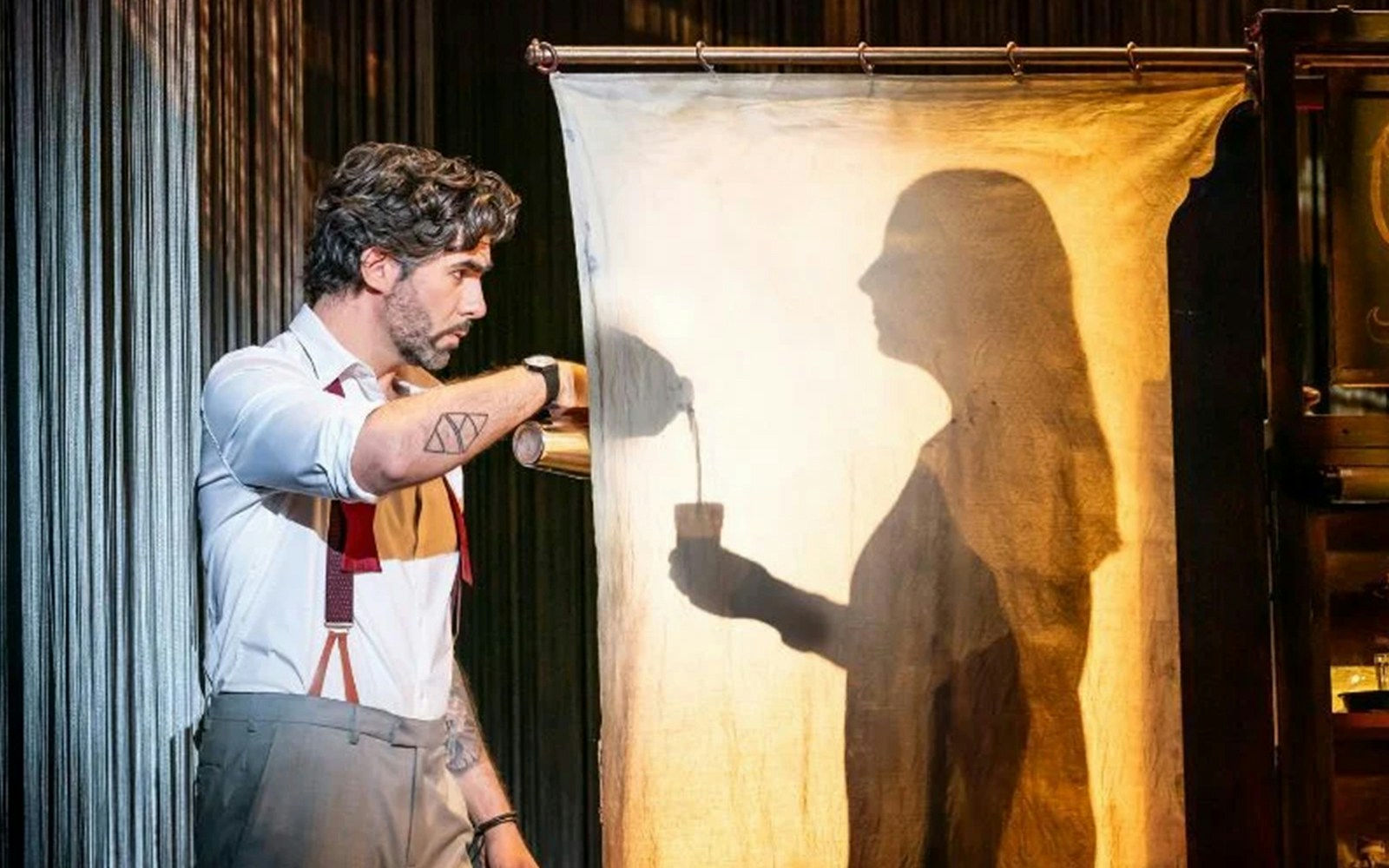 Man pouring drink with shadow silhouette on curtain during theatrical performance.