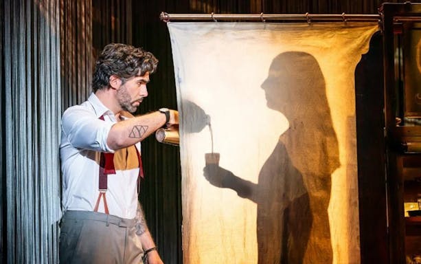 Man pouring drink with shadow silhouette on curtain during theatrical performance.