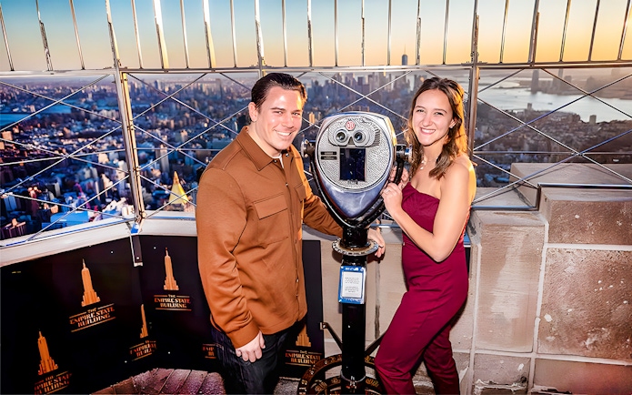 Couple using viewfinder atop Empire State Building, NYC skyline in background.