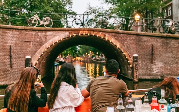 Evening canal cruise in Amsterdam with people enjoying drinks and a view of a lit-up bridge.