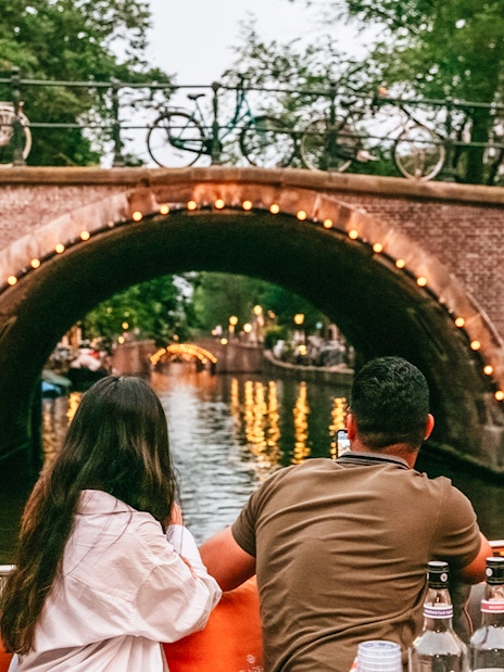 Evening canal cruise in Amsterdam with people enjoying drinks and a view of a lit-up bridge.