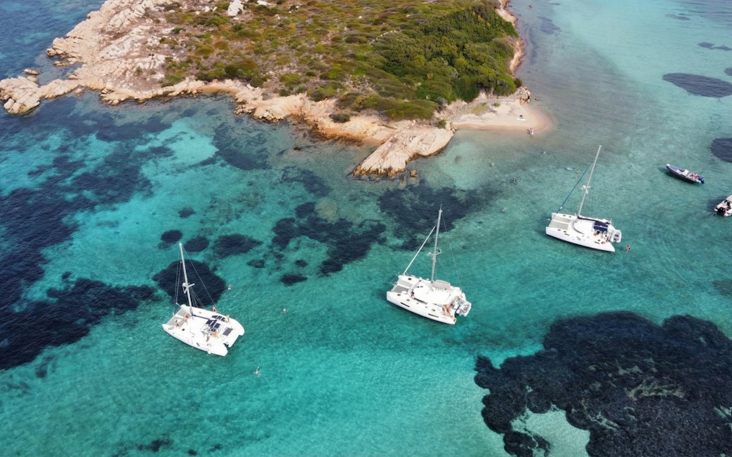 Catamarans sailing near La Maddalena's rocky coastline in clear turquoise waters.
