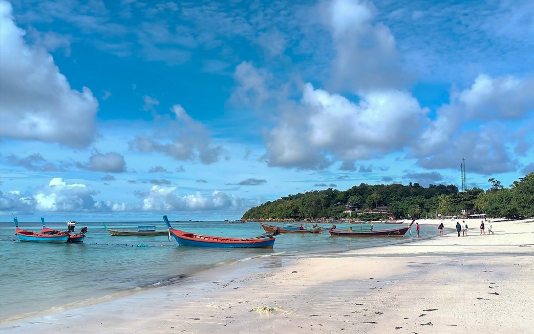 Boats anchored near the shore of Koh Lipe with tourists walking on the beach.