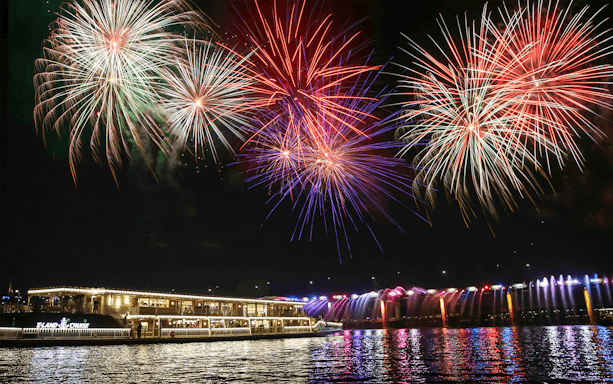 Cruise ship on river with colorful fireworks and illuminated bridge at night.