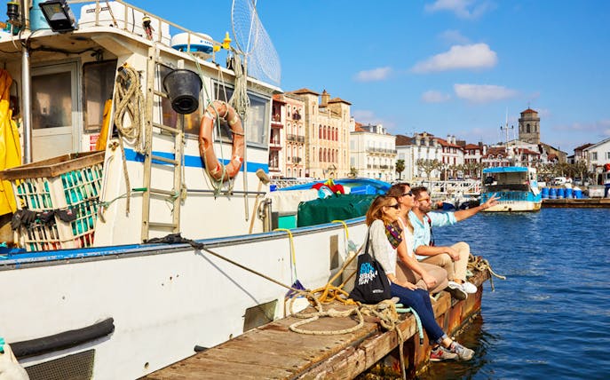 Tourists sitting on a dock by a fishing boat in a French Basque Coast harbor.