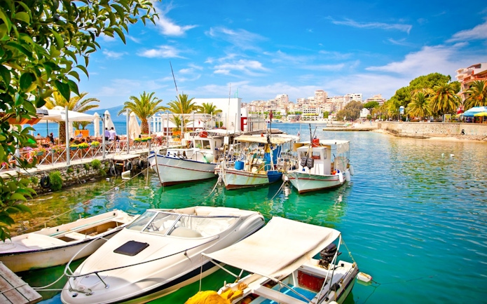Boats docked at Saranda city port, Ionian Sea, Albania, with waterfront cafes and palm trees.