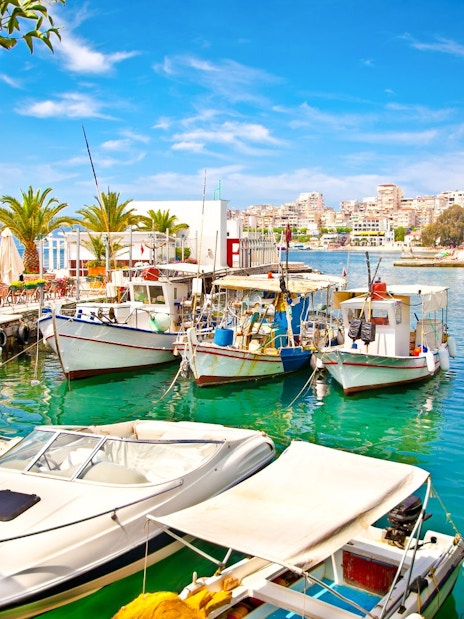 Boats docked at Saranda city port, Ionian Sea, Albania, with waterfront cafes and palm trees.