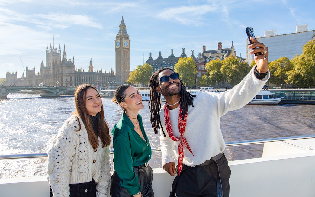 Friends taking a selfie on a Thames River cruise with Big Ben in the background, London.