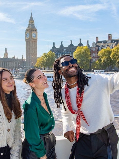 Friends taking a selfie on a Thames River cruise with Big Ben in the background, London.