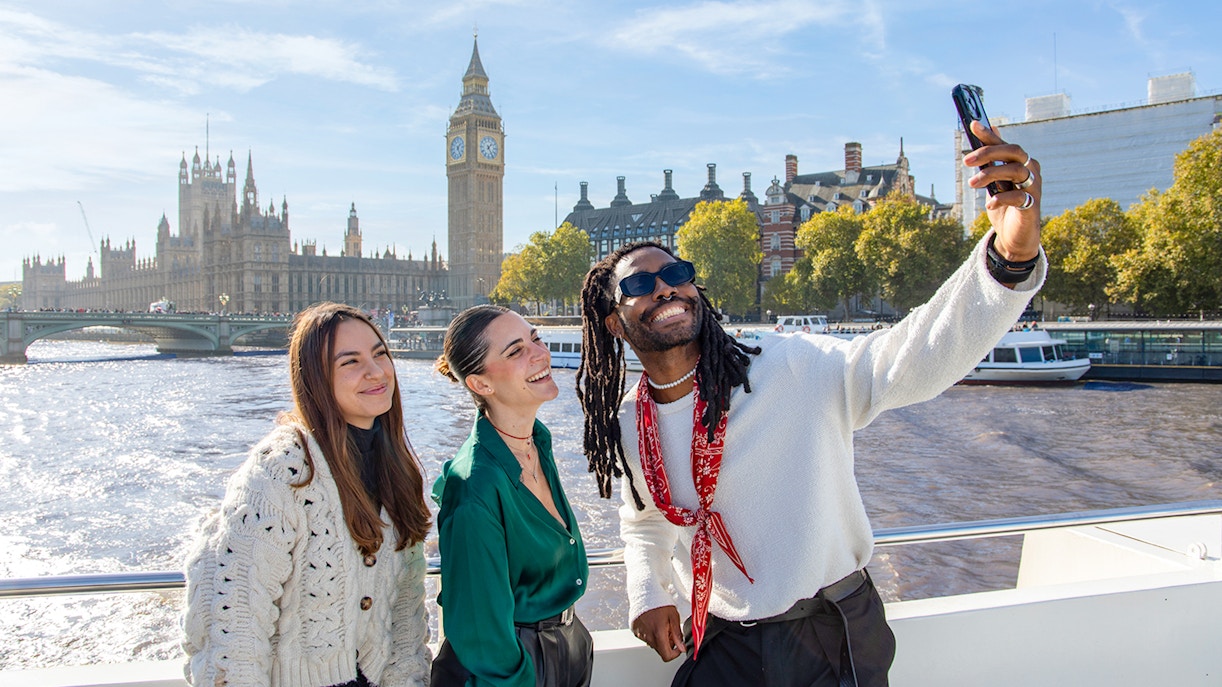 Friends taking a selfie on a Thames River cruise with Big Ben in the background, London.