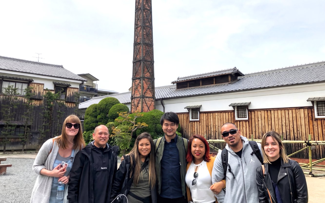 Group of tourists at a Kyoto sake brewery with a tall chimney and traditional wooden buildings.