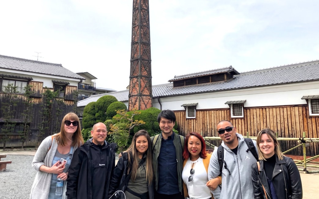 Group of tourists at a Kyoto sake brewery with a tall chimney and traditional wooden buildings.