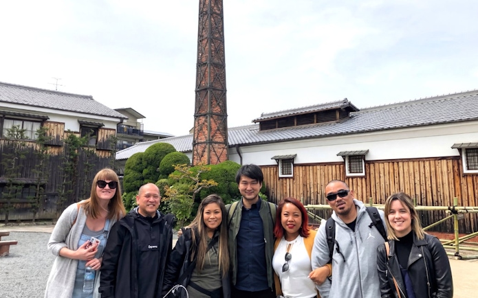 Group of tourists at a Kyoto sake brewery with a tall chimney and traditional wooden buildings.