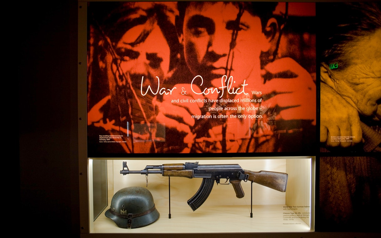 War and Family Migration display with rifle and helmet at Immigration Museum tour.