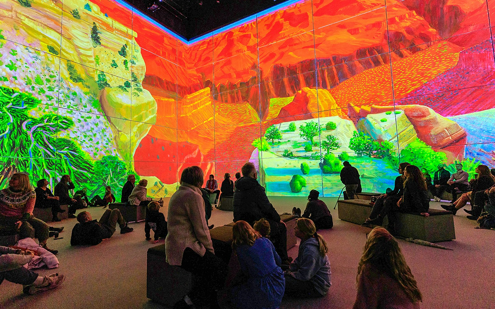 Visitors viewing immersive David Hockney art installation, "Bigger & Closer," with vibrant landscapes.