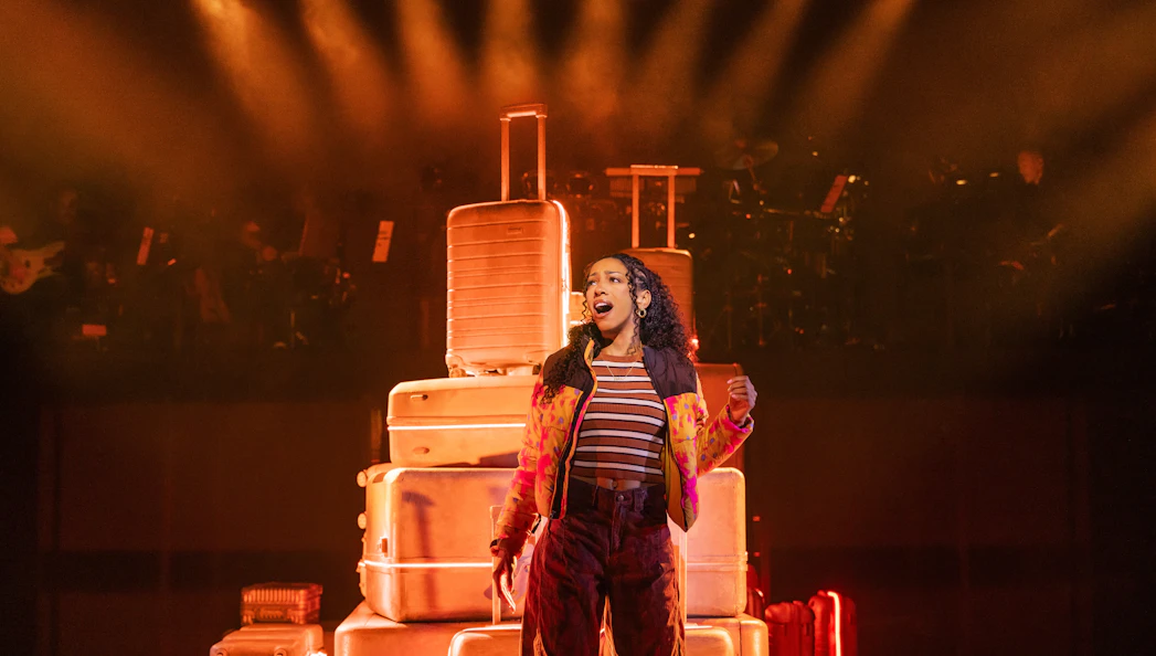 Performer singing on stage with stacked suitcases in "Two Strangers (Carry a Cake Across New York)" Broadway show.