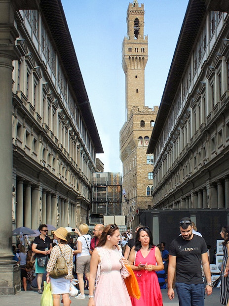 Visitors walking through the Uffizi Gallery courtyard in Florence, Italy, with Palazzo Vecchio in the background.