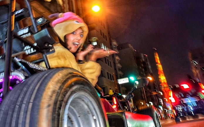Go-karting tour participant in costume driving through Shibuya at night with Tokyo Tower in view.