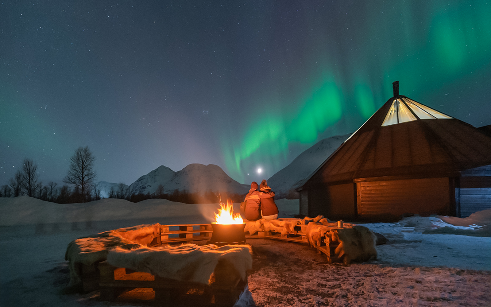Campfire under Northern Lights with snowy mountains and cabin in the background.